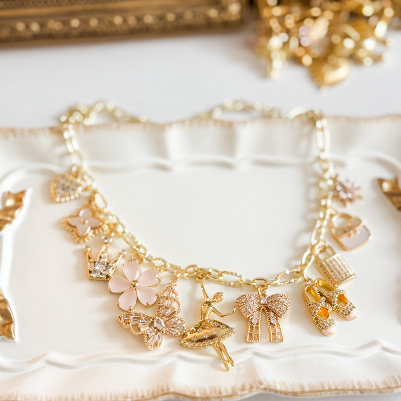 A gold charm bracelet is displayed on a white ceramic tray with scalloped edges. The bracelet features several delicate charms in gold and soft pink tones, including hearts, flowers, a butterfly, a ballerina, a bow, ballet slippers, and sparkling crystal accents. The jewelry is styled in bright, soft lighting with a blurred gold-toned background that creates an elegant, feminine aesthetic.