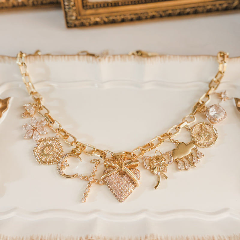 Gold charm necklace displayed on a white decorative tray, featuring a chunky chain adorned with multiple charms including a heart, bow, moon, floral accents, coin medallions, and sparkling gemstone details. The necklace is styled in a flat-lay jewelry photograph with a soft, elegant aesthetic.