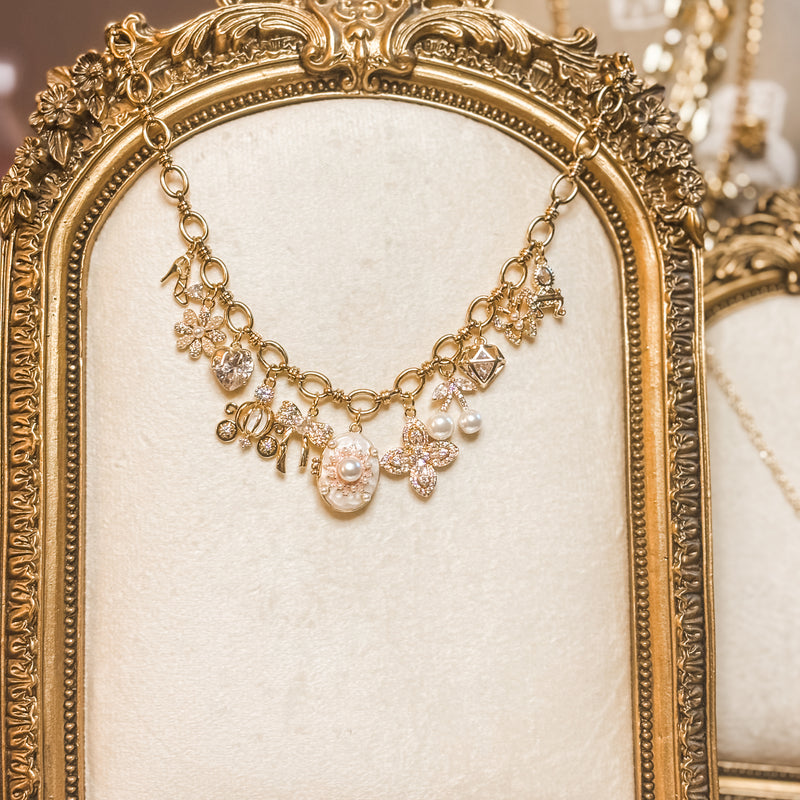 Gold charm necklace displayed on an ornate vintage-style gold frame with a soft ivory velvet backing. The necklace features oval chain links adorned with sparkling charms, including flowers, hearts, bows, pearls, gemstones, and decorative pendants, creating an elegant and feminine design. The warm-toned jewelry photograph is styled with a luxurious, romantic aesthetic.